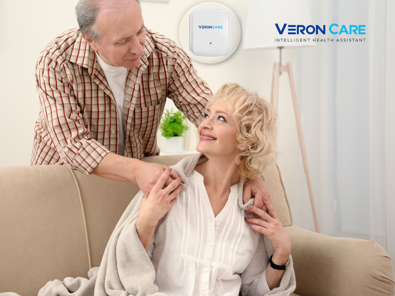 Elderly couple sitting together at home with a Veron Care contactless monitoring device on the wall, symbolizing safe and supportive care.