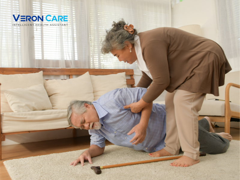 Elderly man on the floor after a fall being helped up by a caregiver in a living room.