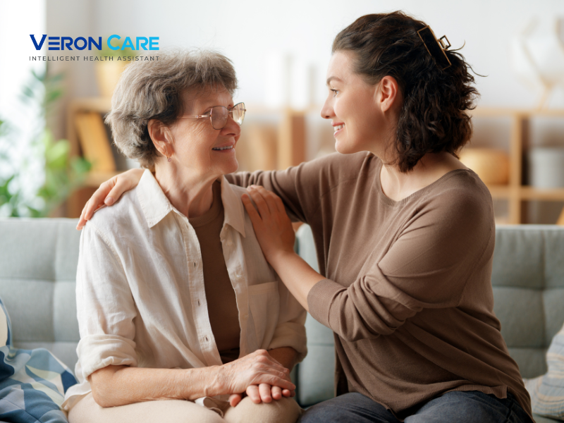 Daughter holding her elderly mother’s shoulders while they smile and talk on a sofa.